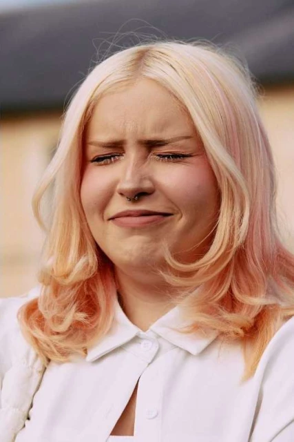 Close-up portrait of an adult female with a slight grimace expression, blonde and peach wavy hair, wearing a white collared shirt.