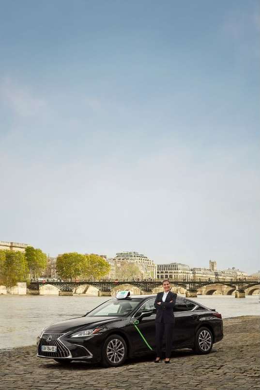 Professional portrait of a woman in a suit standing beside a black Lexus taxi with a scenic river and European architecture background.
