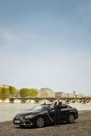 Professional portrait of a woman in a suit standing beside a black Lexus taxi with a scenic river and European architecture background.