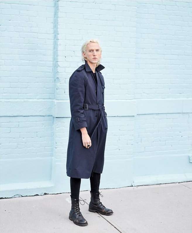 A person with blond hair poses in a dark trench coat, leggings, and boots against a light blue brick wall in an urban setting.