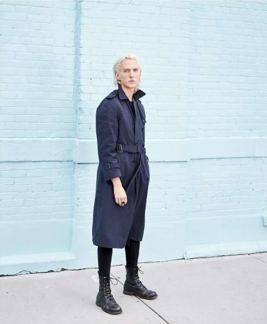 A person with blond hair poses in a dark trench coat, leggings, and boots against a light blue brick wall in an urban setting.