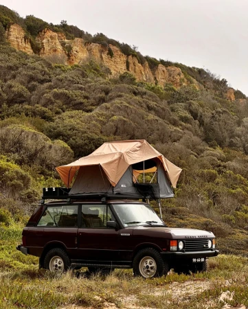 A Range Rover SUV with a rooftop tent parked in a grassy field against a backdrop of coastal cliffs and a cloudy sky.
