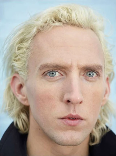 Close-up portrait of a fair-skinned adult male with light blond hair and blue eyes, wearing a dark collared shirt, against a soft blue backdrop.