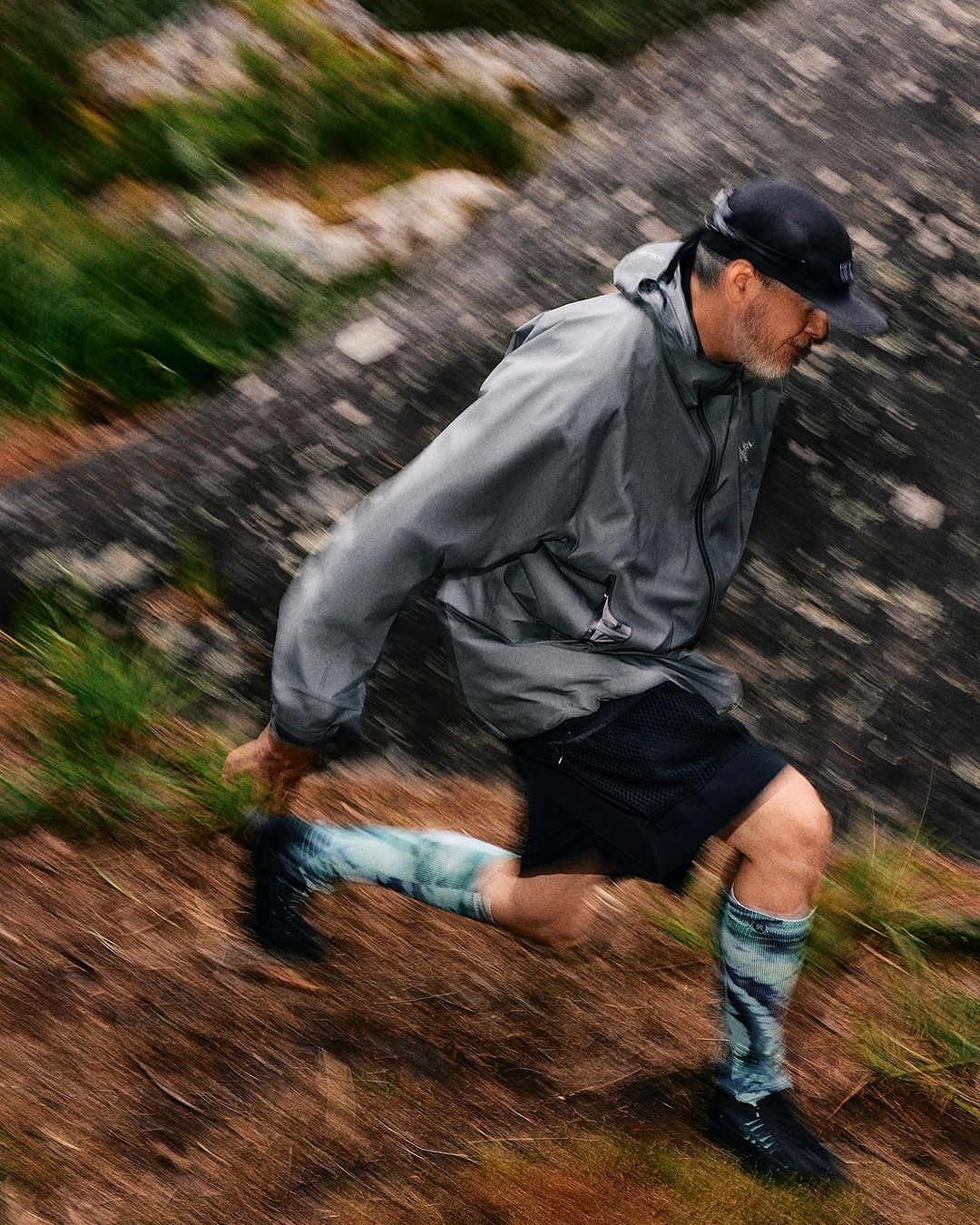 Mature man in athletic gear runs on a grassy slope in an outdoor setting with a large rock formation in the background, captured with motion blur.