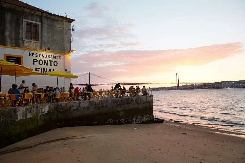 Outdoor restaurant by the water at sunset with people dining under yellow umbrellas, featuring a bridge in the distance against colorful sky.