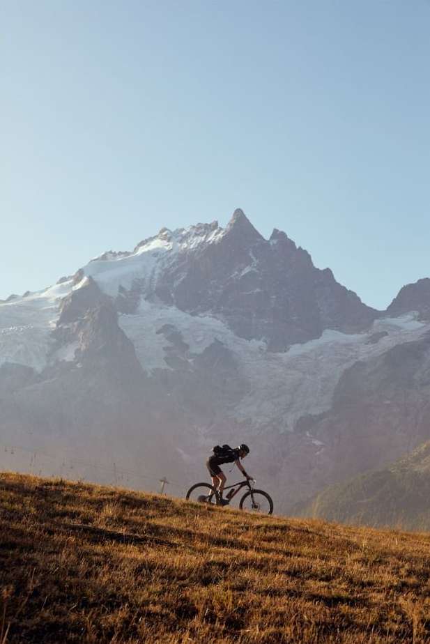 A person rides a mountain bike on a grassy hill, with a view of a large mountain range in the background under a clear sky.
