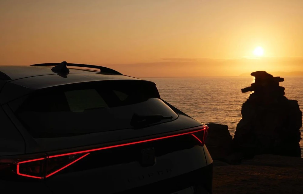 Dark grey Cupra car parked near ocean with rock formations, backlighted by golden sunset. Automotive, outdoors, travel content.