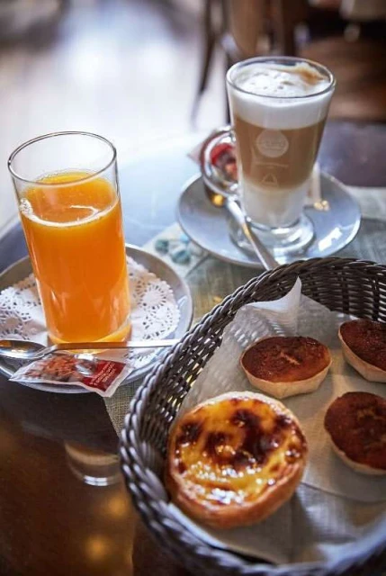 A glass of orange juice, a latte, and a basket of pastel de nata on a table create an inviting food and beverage still life.