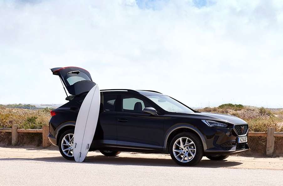 A dark SUV parked on a beach with a surfboard leaning against the open hatchback, under a sunny sky near the ocean.