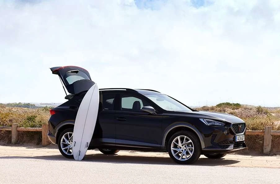 A dark SUV parked on a beach with a surfboard leaning against the open hatchback, under a sunny sky near the ocean.