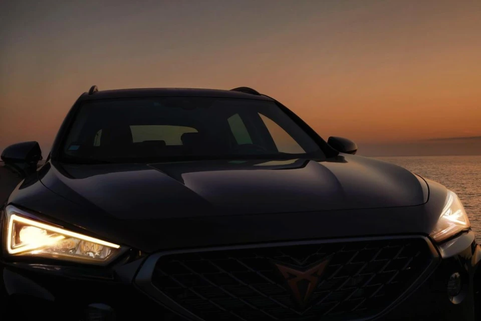 Headlights illuminate a dark car with a black grille against a sunset sky and ocean backdrop, showcasing automotive design.