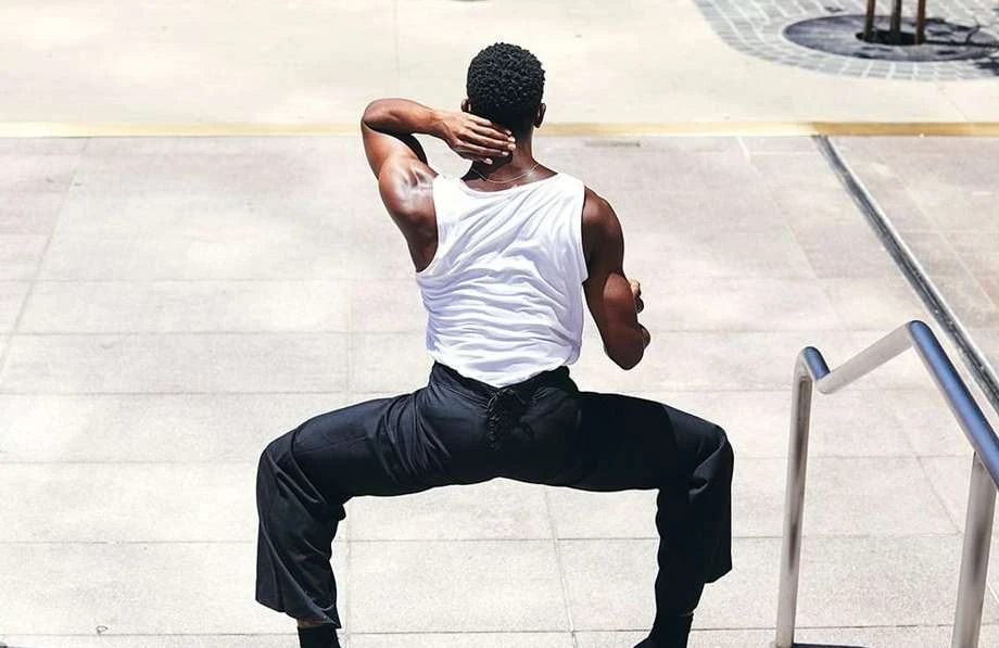 Athletic man in a white tank top and black pants doing martial arts stance on concrete steps with a metal handrail in daylight.