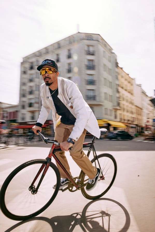 Man rides a fixed-gear bicycle along a street with blurred motion. He wears a jacket, sunglasses, a hat and is partially obscured by motion blur.