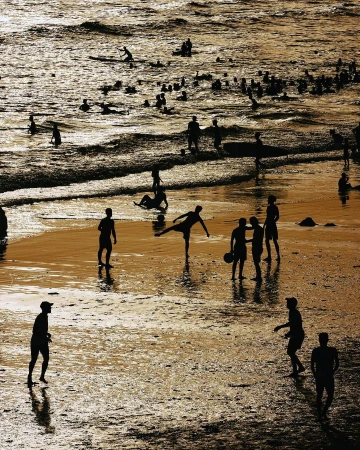 Silhouetted figures play soccer on a sunlit beach with gentle waves and many people in the background, creating a sense of summer leisure.