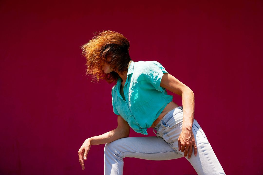 Woman in turquoise blouse and light-wash jeans poses against a bright magenta wall, sunlight illuminating her stance, and the garment details.