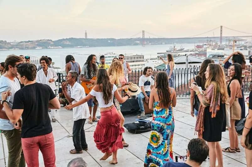 Rooftop party with a diverse group of people enjoying music and dancing with the city and a bridge in the background.