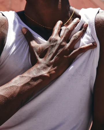 Close-up on a dark-skinned male torso wearing a white tank top, with a hand placed on his chest, and a thin gold chain necklace.