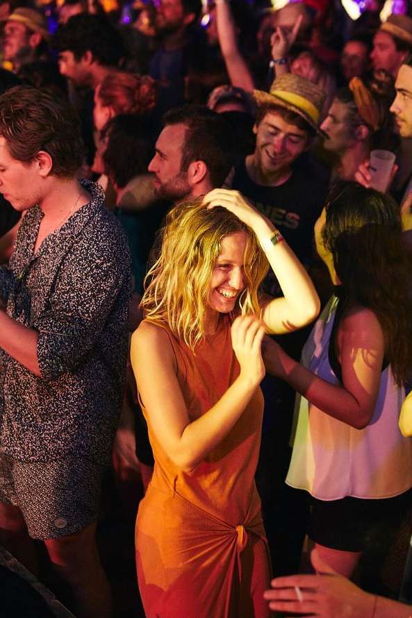 Smiling woman in orange dress dancing in a crowd with other adults, at a music event with dark lighting and a crowded setting.