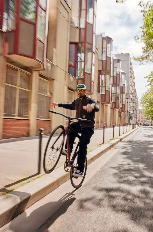 A man on a red bicycle performs a wheelie along a sidewalk in an urban environment with apartment buildings and trees in the background.