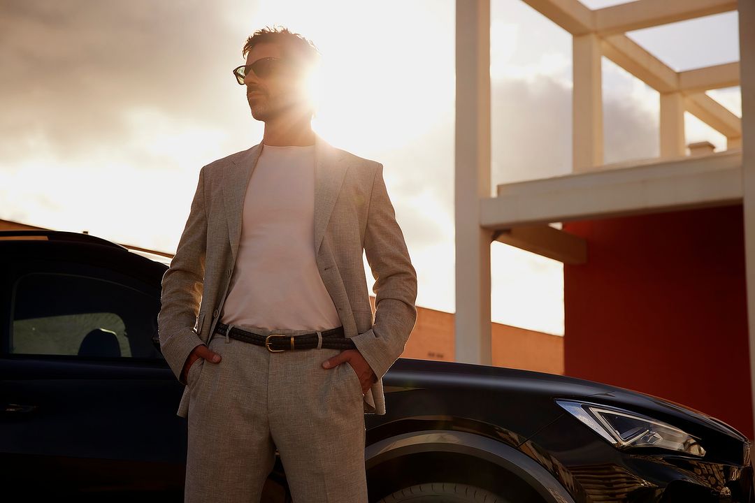 A stylish adult male in a suit poses next to a dark sedan against a bright sky, creating a striking contrast between fashion and automotive.