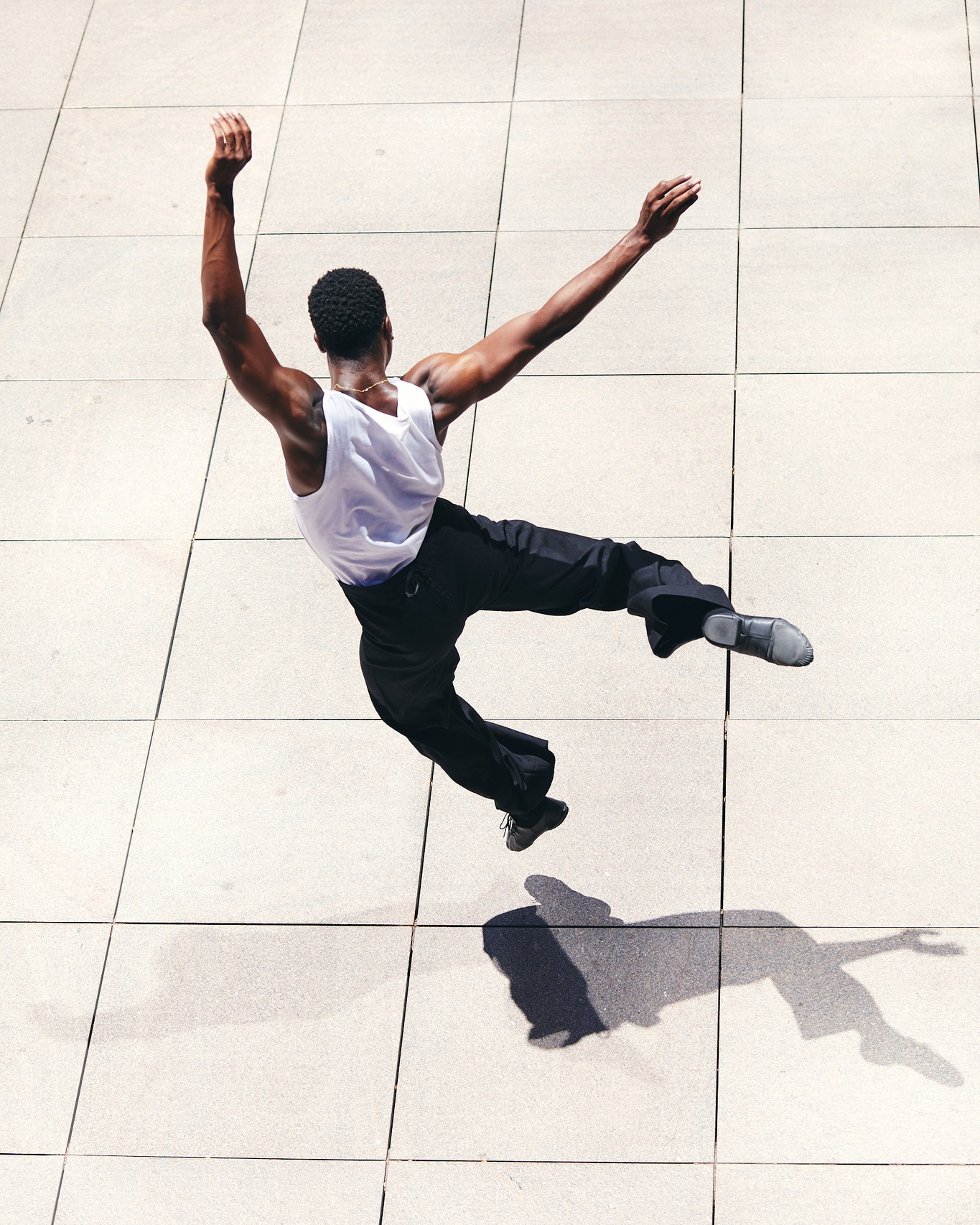 High-angle shot of a muscular Black male dancer in white top and black pants leaping on a tiled floor, casting a shadow below.