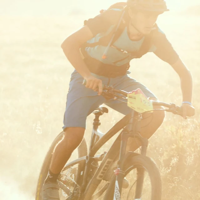 A cyclist wearing blue shorts and a light blue shirt rides a mountain bike on a dirt trail in bright sunlight, creating a sense of outdoor adventure.