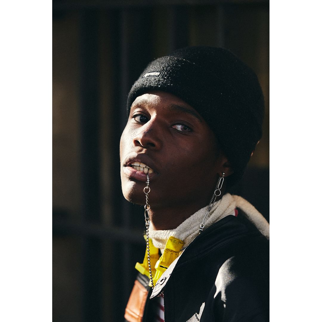 Close-up portrait of a young adult Black man wearing a black beanie and silver jewelry in his lip and ear, looking at the viewer.