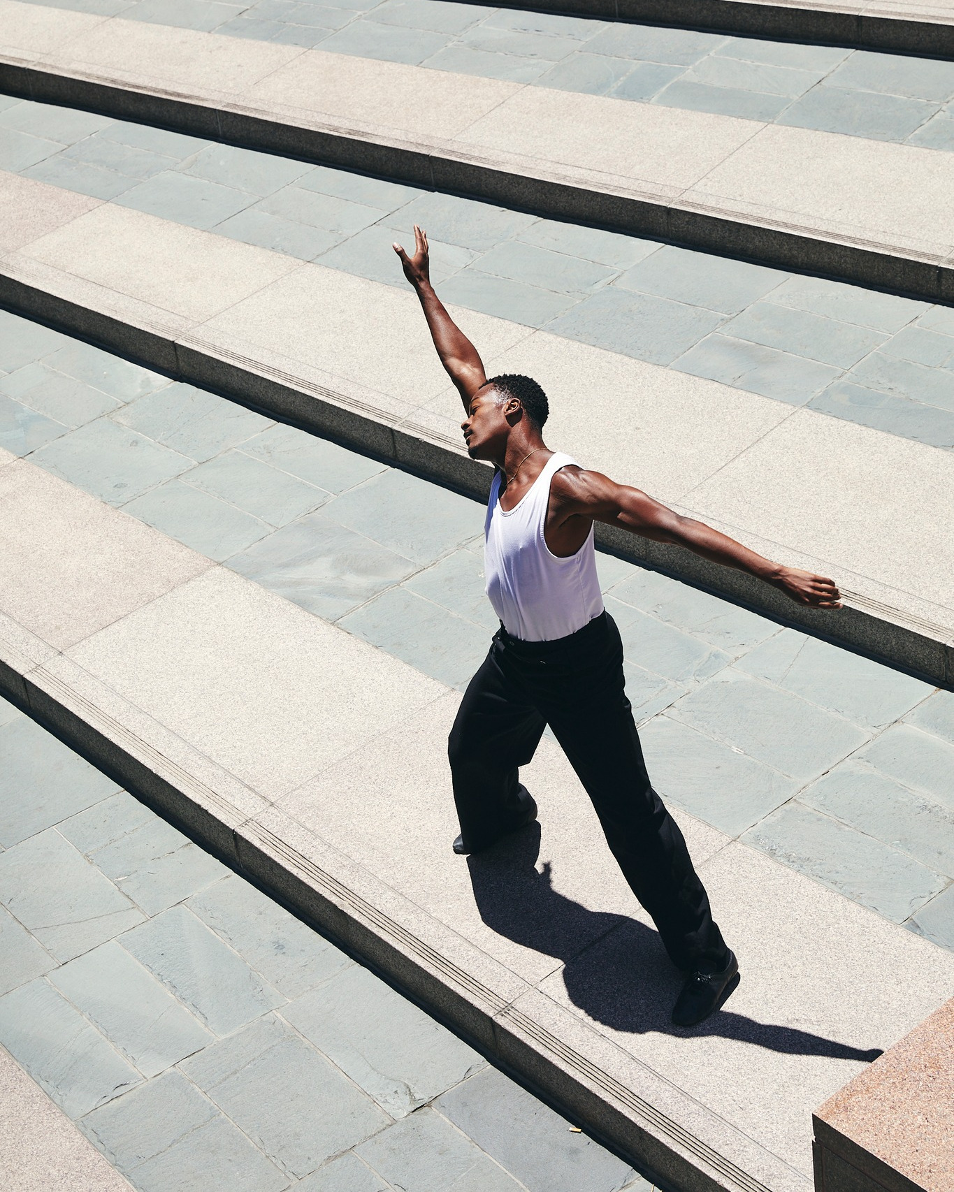 Athletic Black man in white tank top and black pants poses dynamically on concrete stairs outdoors, with bright sunlight casting shadows.