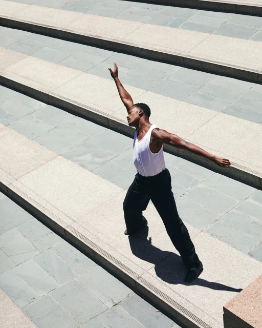Athletic Black man in white tank top and black pants poses dynamically on concrete stairs outdoors, with bright sunlight casting shadows.