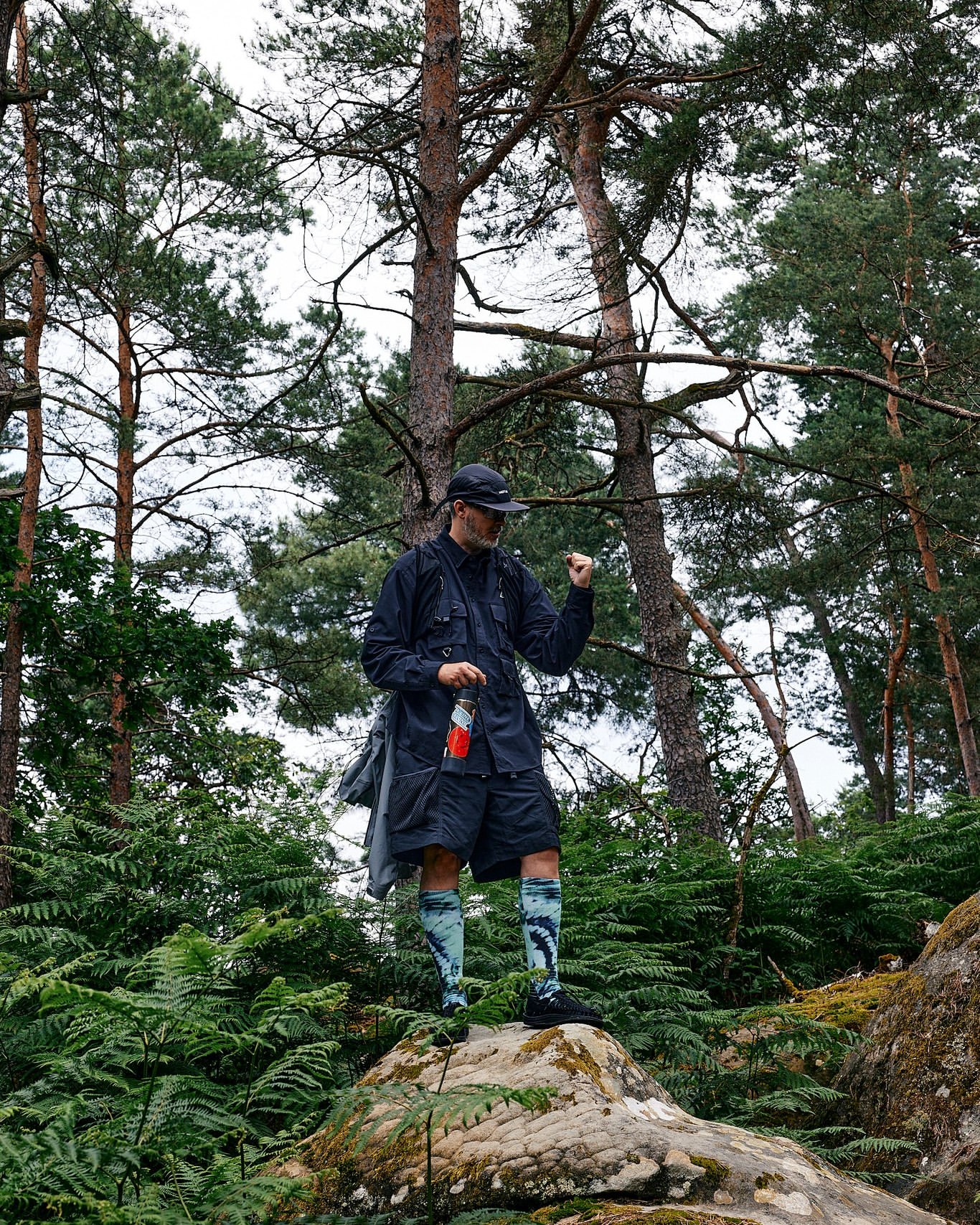 Man in dark outdoor clothing stands on a rock in a forest setting, surrounded by trees, ferns, and wearing patterned socks.