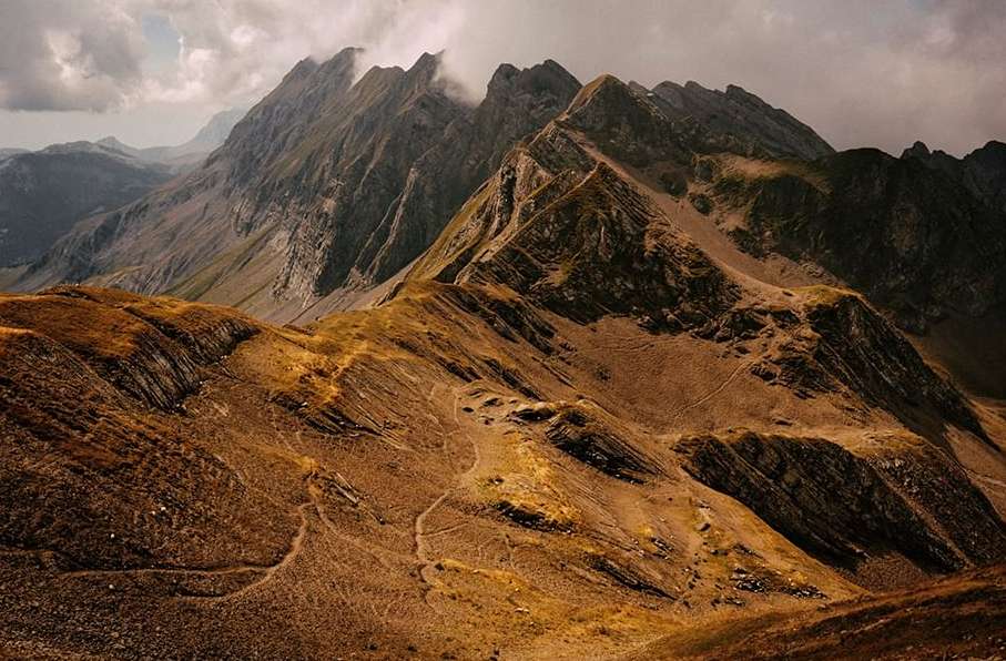 Scenic view of rugged, brown mountains under a cloudy sky, showcasing a vast landscape with winding paths and layered peaks.