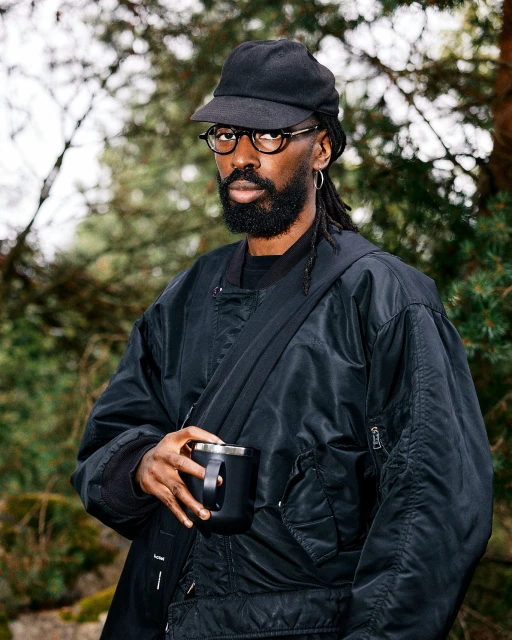 An African-descent adult male in glasses and a cap holds a mug in a forest setting, wearing a black jacket with a crossbody bag.