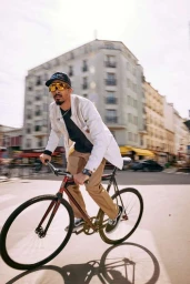 Man wearing a white jacket and gold sunglasses riding a bicycle on a sunny city street in Paris.