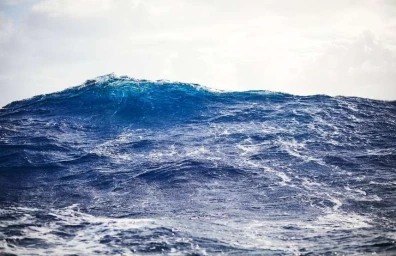 Rolling dark blue ocean wave cresting beneath a cloudy sky, captured in natural daylight by Yoann Stoeckel.