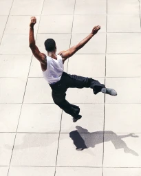 High angle shot of a Black man mid-air jumping outdoors over light tiled ground wearing a white tank top and black trousers