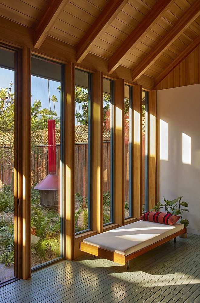 Indoor sunroom with floor-to-ceiling windows overlooking a landscaped yard, a wooden daybed with a striped cushion, and a wood-paneled ceiling.