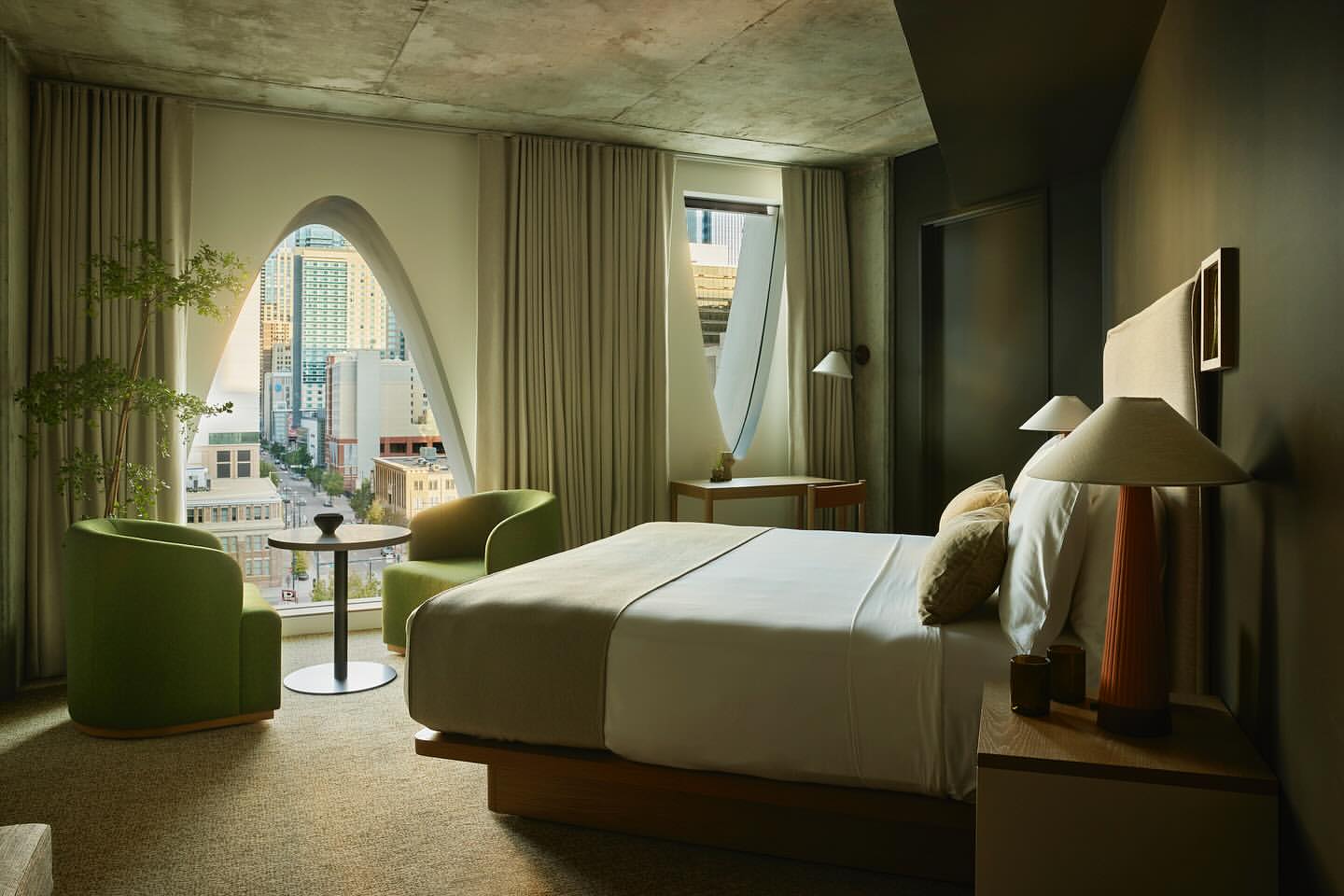 Modern hotel room interior featuring a bed, seating area with chairs and table, and a cityscape view through a unique arched window.