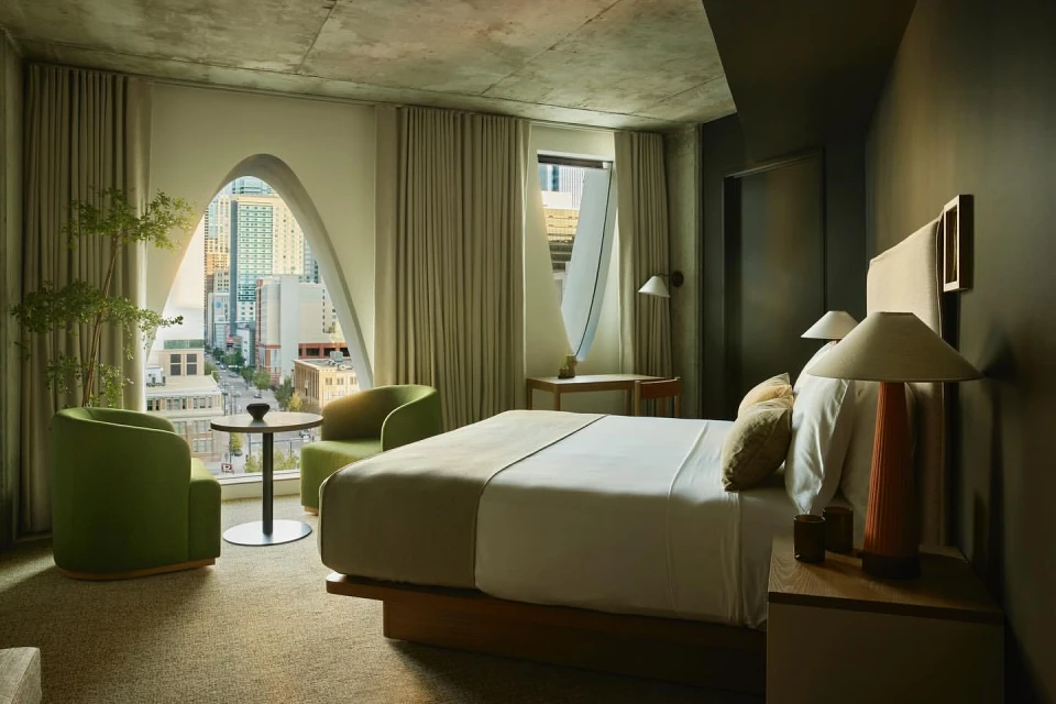 Modern hotel room interior featuring a bed, seating area with chairs and table, and a cityscape view through a unique arched window.