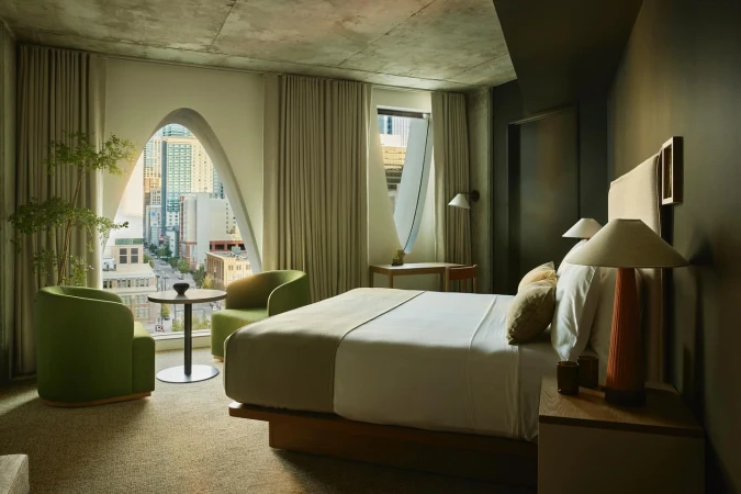 Modern hotel room interior featuring a bed, seating area with chairs and table, and a cityscape view through a unique arched window.