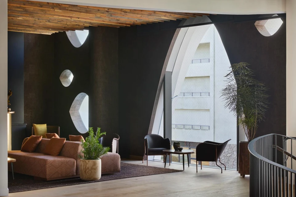 Contemporary living room interior with a brown sofa, dark armchairs, a round table, potted plants, and large windows offering city views.