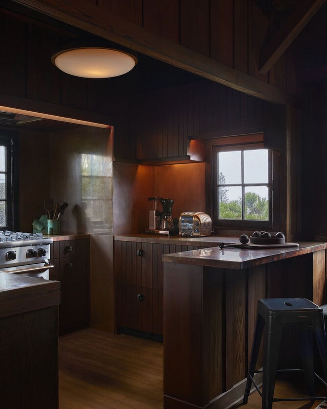 Dark brown kitchen interior featuring wood cabinets, countertop appliances, bar stool, under-cabinet lighting, and window with view.