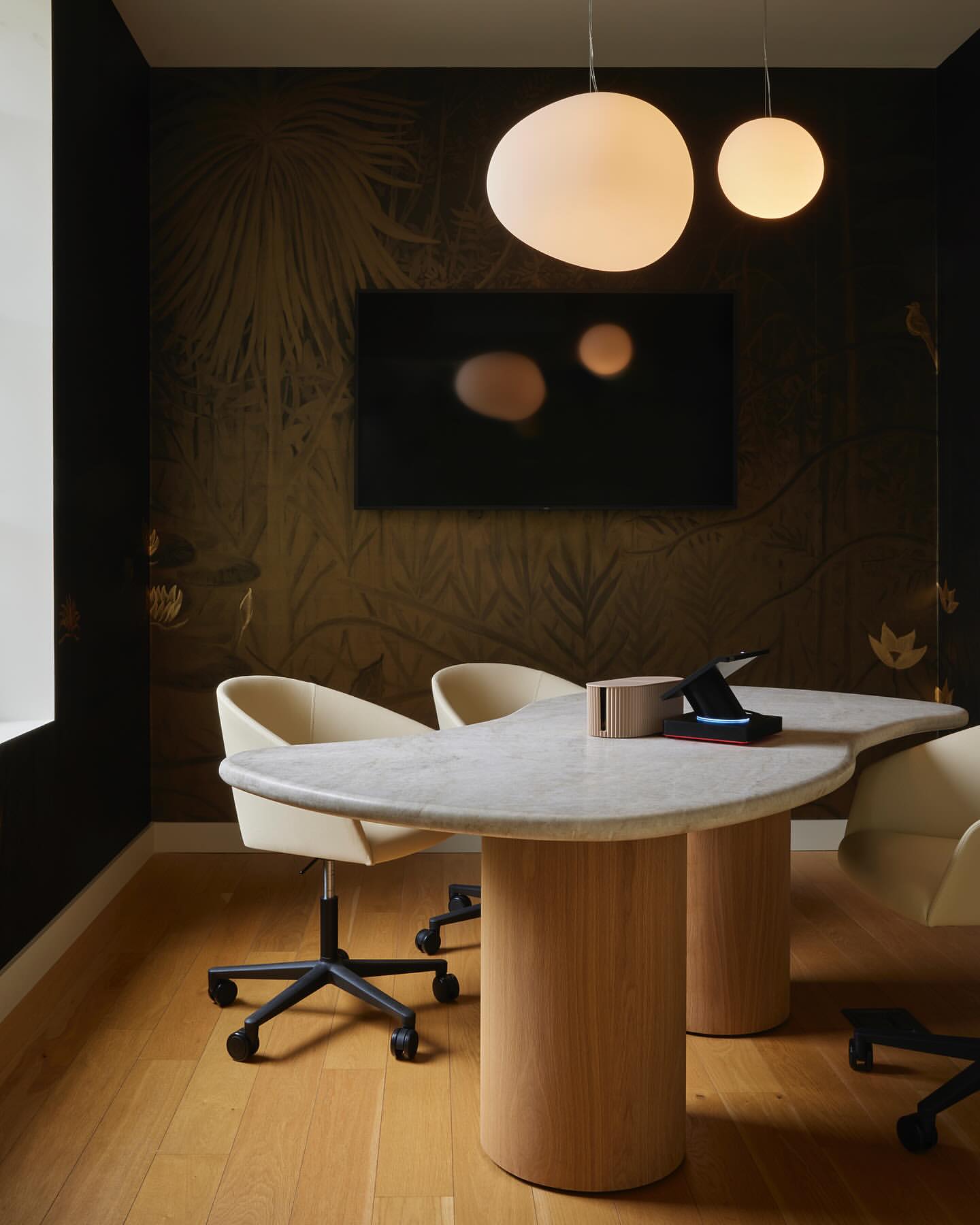 Conference room interior with marble table, round pendant lights, and stylish office chairs against a botanical wallpaper backdrop.