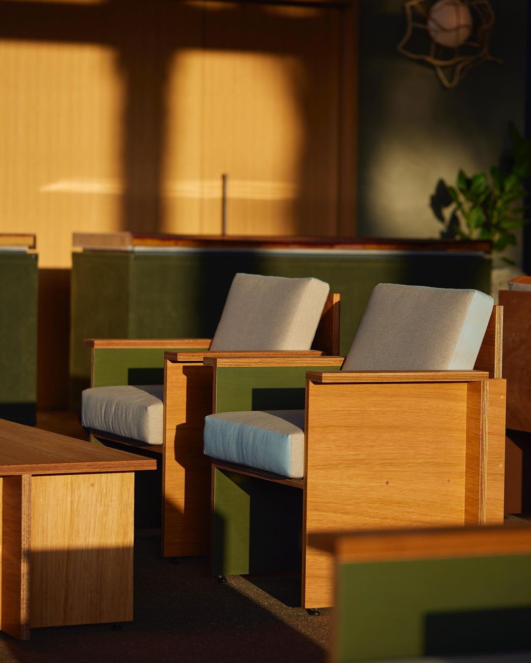 Interior scene with two wooden armchairs with cream cushions, placed around a small table; warm light from the side.