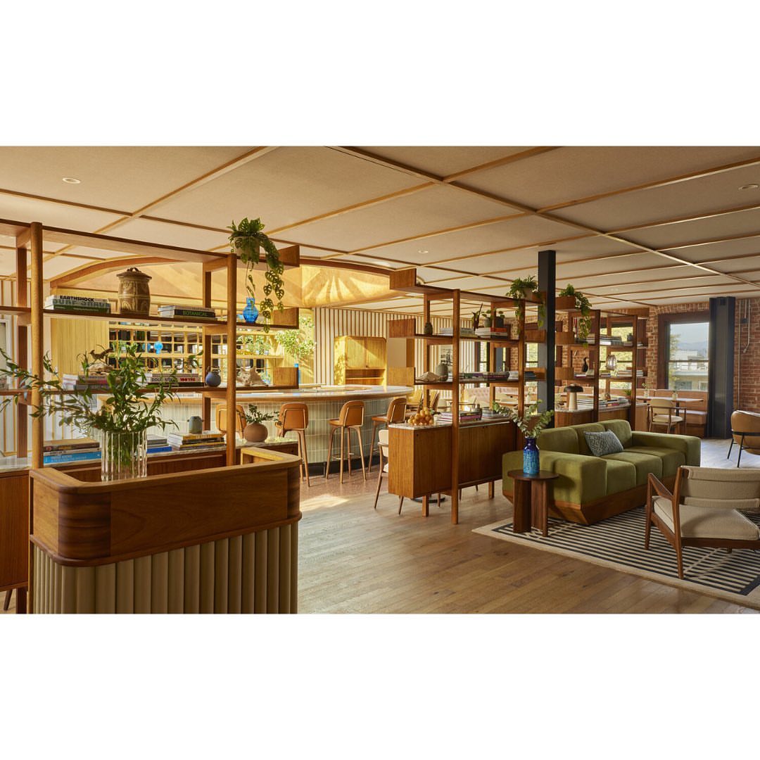 Interior of a hotel lobby featuring mid-century modern furniture, open shelving, plants, and wood flooring with natural light from windows.