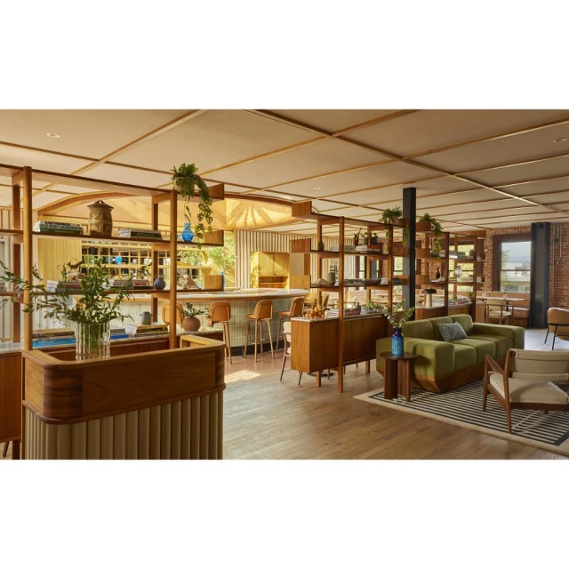 Interior of a hotel lobby featuring mid-century modern furniture, open shelving, plants, and wood flooring with natural light from windows.