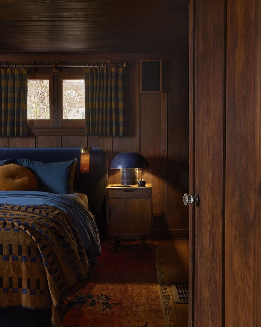 Cozy bedroom interior with a bed covered in a patterned blanket, a bedside table with a lamp, and wooden walls, creating a warm ambiance.