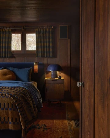Cozy bedroom interior with a bed covered in a patterned blanket, a bedside table with a lamp, and wooden walls, creating a warm ambiance.