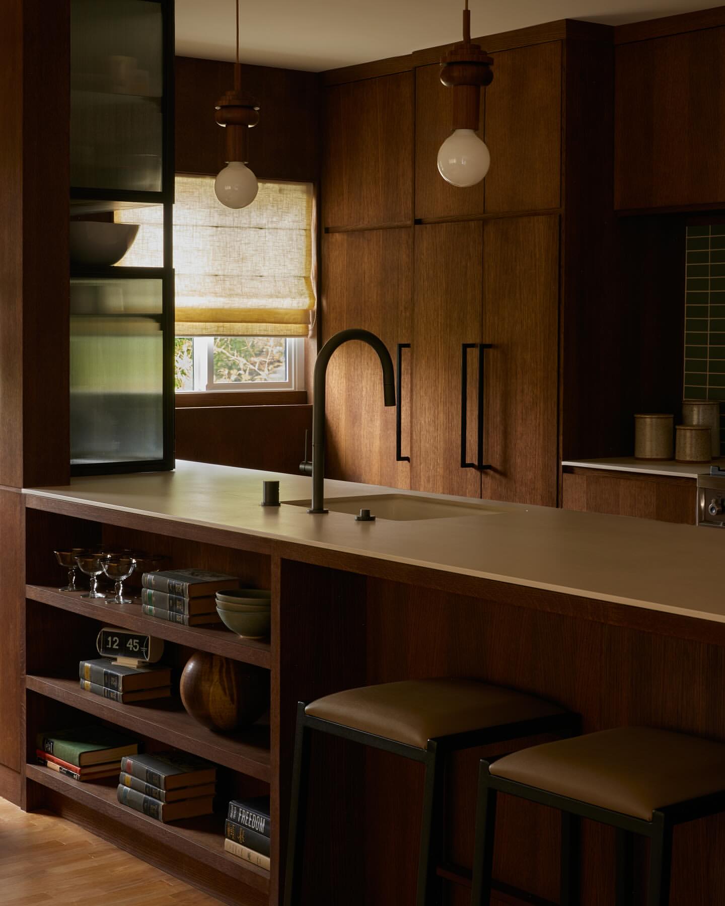 A warm-toned kitchen interior featuring dark wood cabinets, a light countertop, a black faucet, two stools, and soft, ambient lighting.