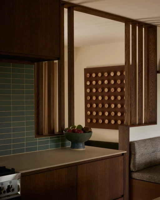 Modern kitchen interior featuring brown wood cabinetry, light countertop, green tiled backsplash, and a bowl of fruit on the counter.