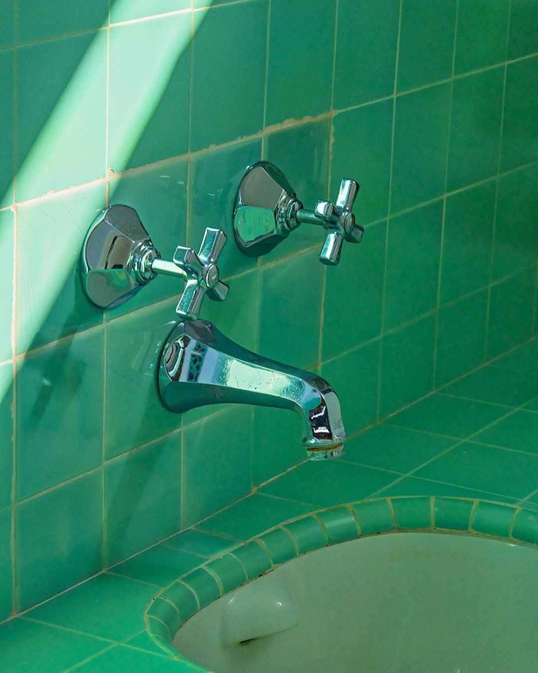 Close-up of a chrome faucet with cross handles mounted on a green tiled wall and sink edge, with bright light casting shadows.